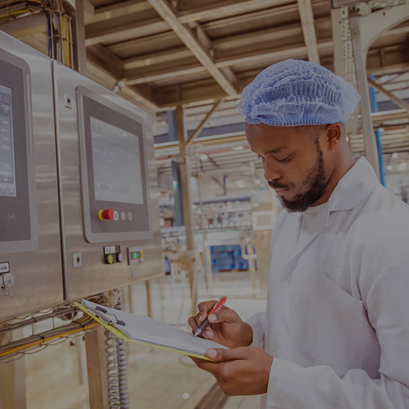 man in manufacturing facility in white coat and hair cap writing on clipboard in front of equipment